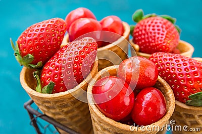 Ripe organic strawberries, glossy sweet cherries in waffle ice cream cones in wire basket, blue background, styled image Stock Photo