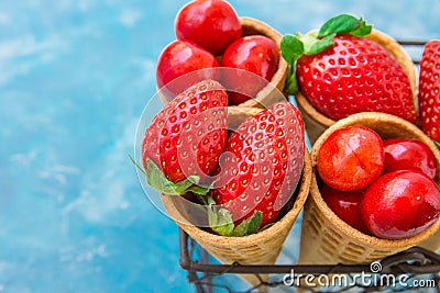 Ripe organic strawberries, glossy sweet cherries in waffle ice cream cones in wire basket, light blue background Stock Photo