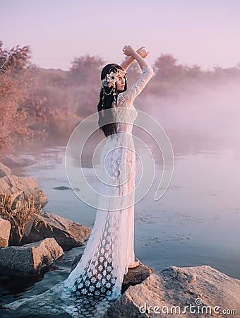 A river nymph in a white lace dress stands on a rock by the lake. The princess has a beautiful wreath with seashells Stock Photo