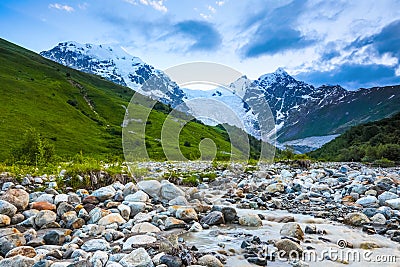 From the river shore, covered with stones, opens view on fantastic glacier and steep rocky mountains with green meadows. Stock Photo