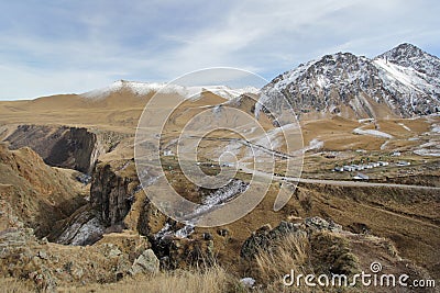 A road and tiny houses against the backdrop of yellow grass, snow-capped mountain peaks and rocky cliffs Stock Photo