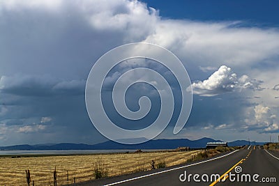 Road trip - Black top two lane highway stretches over the horizon past a barn and hay field by a lake towards the distant mountain Stock Photo