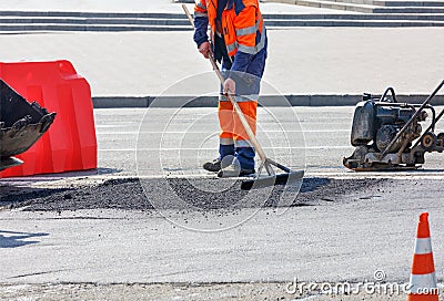 A road worker in a bright orange overalls levels fresh asphalt with a wooden level for later compaction Stock Photo