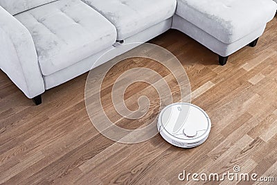 Robotic vacuum cleaner runs near sofa on laminate floor. Robot controlled by voice commands to direct cleaning. Modern smart Stock Photo