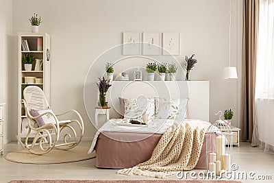 Rocking chair next to bed with blanket in provencal bedroom interior with plants and posters. Real photo Stock Photo