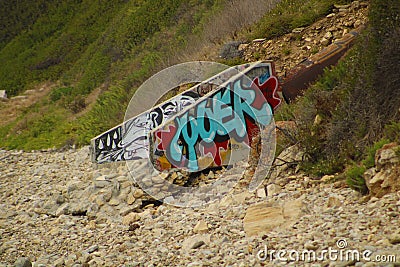 A rocky beach with a lush green hillside and graffiti painted on the walls of the storm drain Editorial Stock Photo