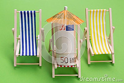 A roll of euro notes lies in a lounge chair, under an umbrella, as if resting, beside it stand two empty lounge chairs Stock Photo