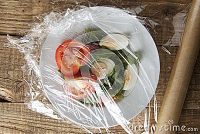 Roll of wrapping plastic stretch film over plate of vegetabes. Close-up isolated on wooden background. No plastic concept. Zero we Stock Photo