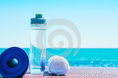 Rolled Yoga Mat Bottle with Water White Towel on Beach with Turquoise Sea Blue Sky in Background. Sunlight. Relaxation Meditation Stock Photo