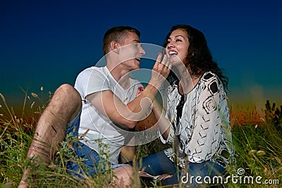 Romantic couple eating red raspberry, sit on grass at sunset on outdoor, dark night sky, love concept, young adult people Stock Photo