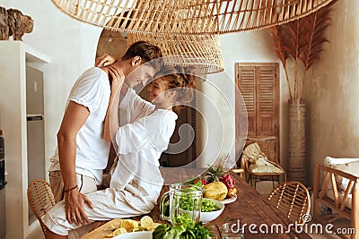 Romantic Couple At Kitchen. Beautiful Woman Sitting On Wooden Table And Hugging Standing Man. Stock Photo