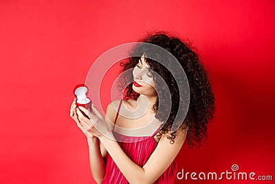 Romantic curly-haired woman in red dress, looking happy at engagement ring, become a bride, standing on studio Stock Photo