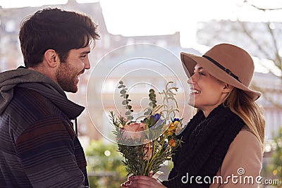 Romantic Man Surprising Young Woman With Bouquet Of Flowers As They Meet In City Park Stock Photo