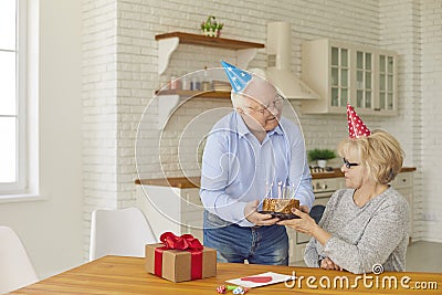 Romantic older man gives a cake to his wife to make a wish and blow out the candles. Senior couple in colored cone hats Stock Photo