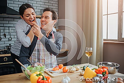 Romantic picture of man feeding woman with piece of vegetable. They are together in kitchen. He sit at table while she Stock Photo