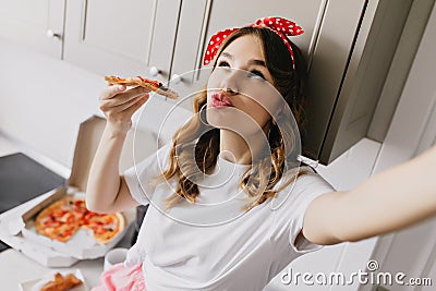 Romantic white girl making selfie while eating pizza. Indoor shot of curly caucasian lady fooling a Stock Photo