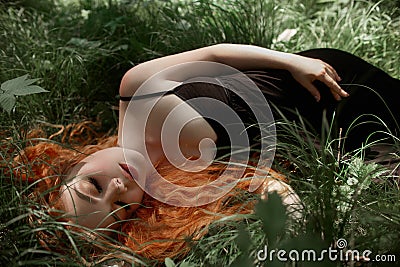 Romantic woman with red hair lying in the grass in the woods. A girl in a light black dress sleeps and dreams in a magical forest. Stock Photo