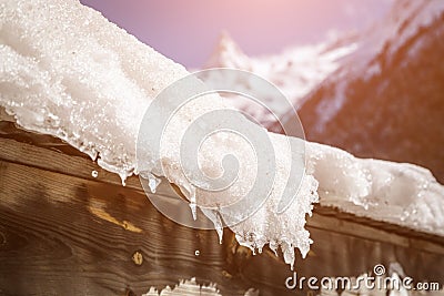 Roof of the house is covered with a thick layer of snow and icicles on the background of snow-capped mountains and bright sun Stock Photo