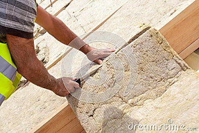 Roofer builder worker installing roof insulation material on new house under construction. Cutting rockwall with sharp knife. Stock Photo