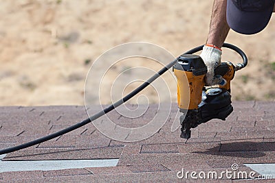 Roofer builder worker with nailgun installing Asphalt Shingles or Bitumen Tiles on a new house under construction. Stock Photo