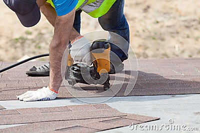 Roofer builder worker with nailgun installing Asphalt Shingles or Bitumen Tiles on a new house under construction. Stock Photo
