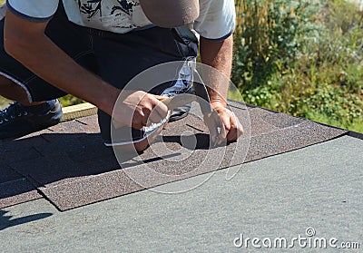 Roofer with hammer and nails laying asphalt shingles tiles on house roof. Roofing Construction Stock Photo