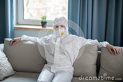 Woman in white workwear and respirator sitting on the sofa Stock Photo