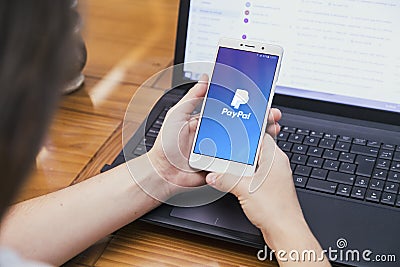 Paypal on the screen of smartphone.Girl in front of a laptop with cell phone. Editorial Stock Photo