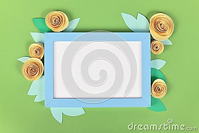 Rose flat lay with blue empty picture frame surrounded by yellow romantic paper craft flowers on green background Stock Photo