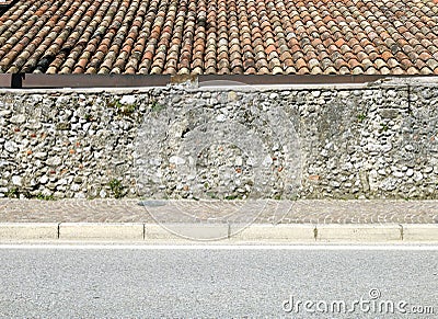 Rough wall made of concrete and stones with a tiled rooftop behind it. Sidewalk and asphalt road in front. Stock Photo