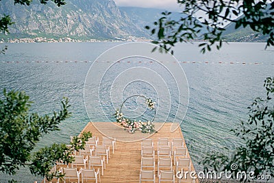 Round wedding arch stands on a wooden pier in front of rows of white chairs Stock Photo