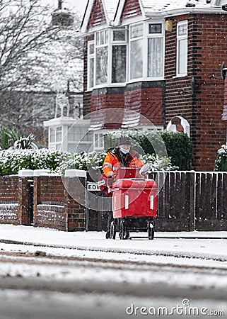 Royal Mail British post man in deep cold snow falling on road and icy frozen pavement delivering letters with trolley red uniform Editorial Stock Photo