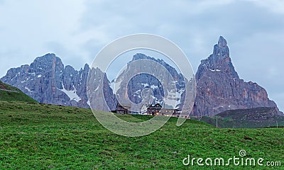 Rugged Cimon della Pala among peaks of Pale di San Martino mountains with a hotel on green grassy hill in Passo Rolle Stock Photo