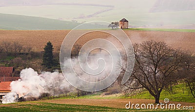 Rural Pastoral Spring Landscape With Old Windmill Without Blades And Plowed Land On Background Of The Village And Thick White Smok Stock Photo