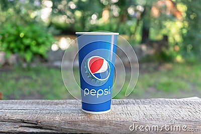 08/05/2020 Russia. Saint Petersburg. A disposable glass with the Pepsi Cola logo stands on a wooden tabletop, on a summer green ba Editorial Stock Photo