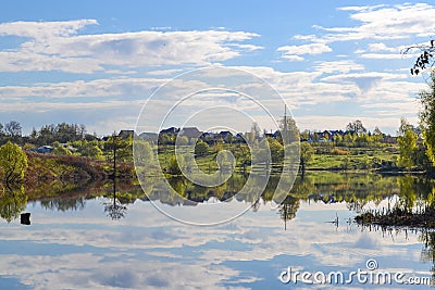 Russian village and green fields with trees and a blue sky with clouds reflected in the calm water of the lake. Calm and silence. Stock Photo