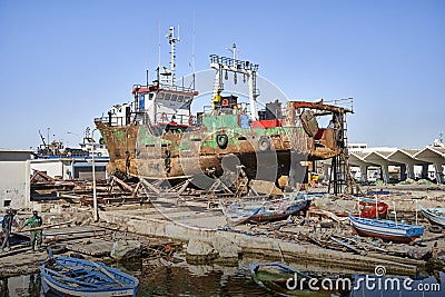 Rusted ship with only a few remains of paint on the iron hull stands in the dry dock in front of the small boats of the fishermen Editorial Stock Photo