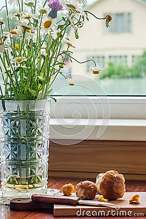 Rustic scene - old-fashionned glass vase with bouquet of chamomiles and wooden desk with mashrooms and knife on window background Stock Photo
