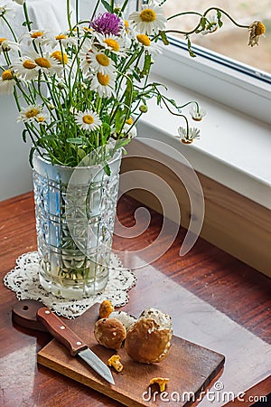 Rustic scene - old-fashionned glass vase with bouquet of chamomiles and wooden desk with mashrooms and knife on window background Stock Photo