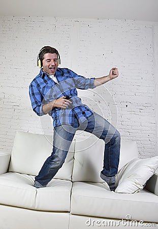 20s or 30s man jumped on couch listening to music on mobile phone with headphones playing air guitar Stock Photo