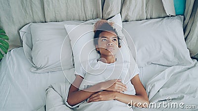 Sad African American lady is lying in bed and sighing looking at ceiling. Comfortable bed, pillows and sheets are Stock Photo