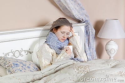 Sad alone young woman in white sweater and blue scarf feeling cold sick and resting home in bed. holding her hot drink and try to Stock Photo