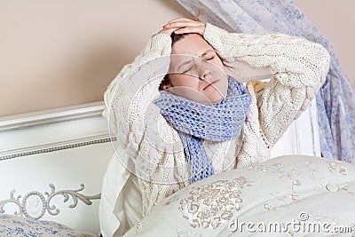 Sad alone young woman in white sweater and blue scarf feeling cold sick and resting home in bed. holding her painful head and Stock Photo