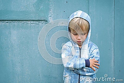 Sad bored resentful child boy in hoodie standing near blue wall. Cute upset distressed kid pouts his lips. Emotional portrait Stock Photo