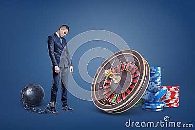 A sad businessman chained to an iron ball stands near a casino roulette and chip stacks. Stock Photo