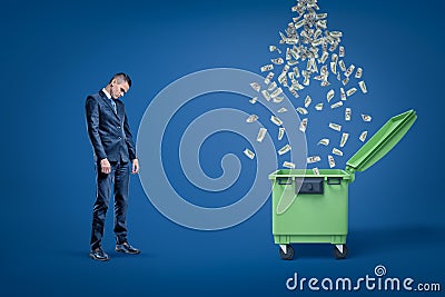 Sad businessman looking down standing next to open dumspter with rain of dollars falling into it. Stock Photo