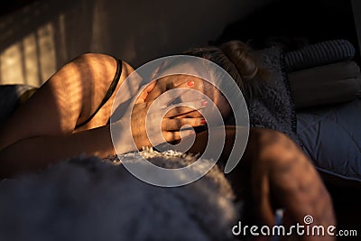 Sad, depressed blonde woman lying alone in bed, face covered with hands, with morning light Stock Photo