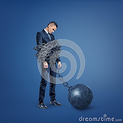 A sad disappointed businessman stands bound by a wide metal chain which is locked to an iron ball. Stock Photo