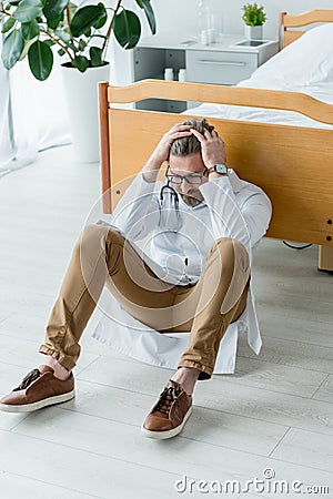 sad doctor in white coat sitting on floor and touching head in hospital. Stock Photo