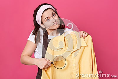 Sad female wears white t shirt, brown apron and hair band, holds yellow blouse and magnifier in front of large, looks at camera Stock Photo